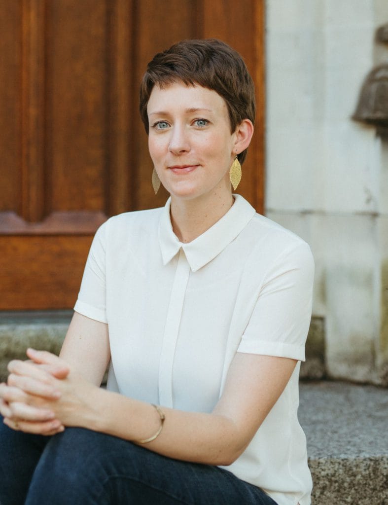 A white woman in her 30s with a brown pixie wearing a white button down shirt and blue jeans sitting on church steps. 