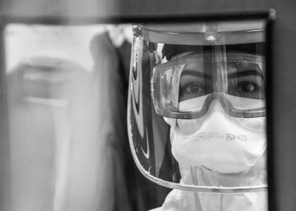 A healthcare worker in full PPE looking through a glass door with an overwhelmed expression.
